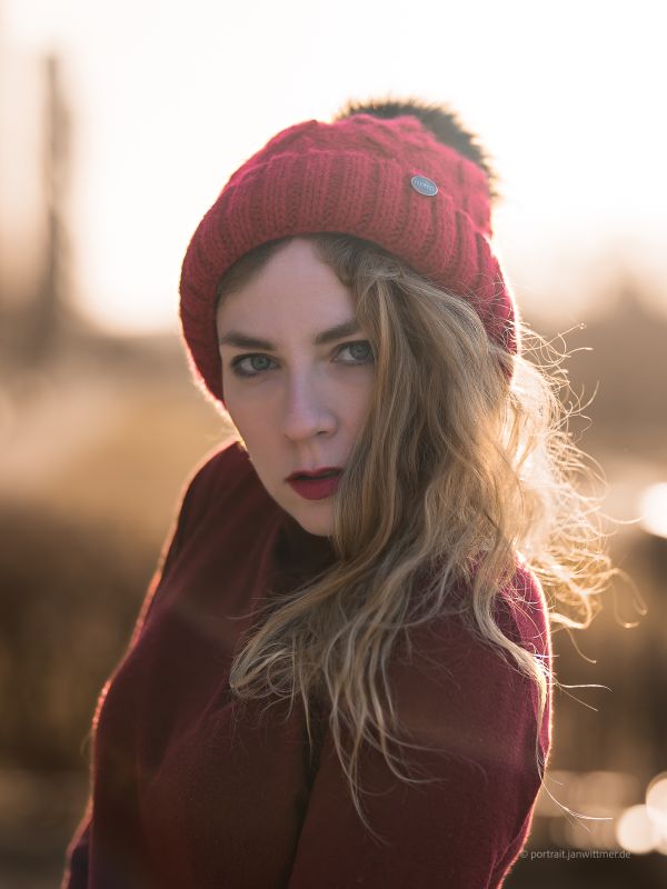 Frau mit roter Bommelmütze und weinrotem Pullover im winterlichen Gegenlicht blickt in die Kamera
