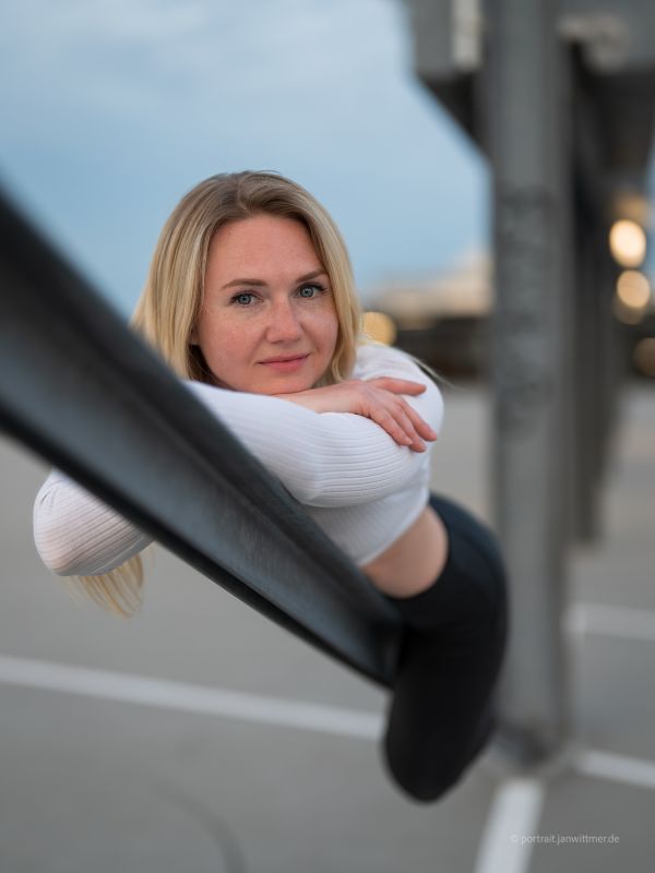 Blonde Frau in weißem Rippshirt lehnt sich auf einem Parkdeck über ein Geländer, blaue Stunde