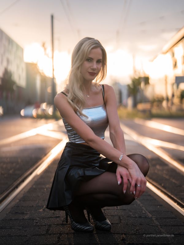 Blonde Frau in silbernem Top und schwarzem Rock hockt auf Straßenbahnschienen im Gegenlicht der tiefen Sonne